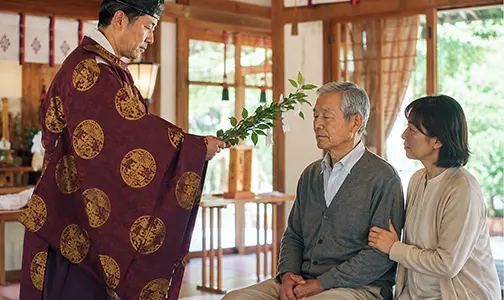 神社でのご祈祷で、神職から玉串を受け取るシニア男性と、その様子を隣で見守る女性