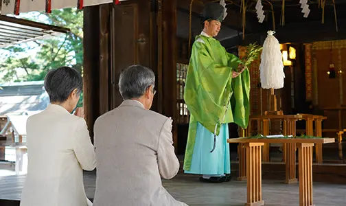 神社の本殿にて、緑色の装束を着た神職によるご祈祷を、後ろ姿で手を合わせながら受けるシニアの参拝客