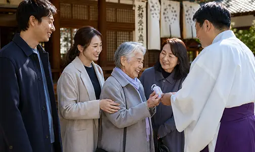 神社境内にて、神職からお守りを受け取り笑顔を見せる高齢の女性と、それを見守る家族の様子